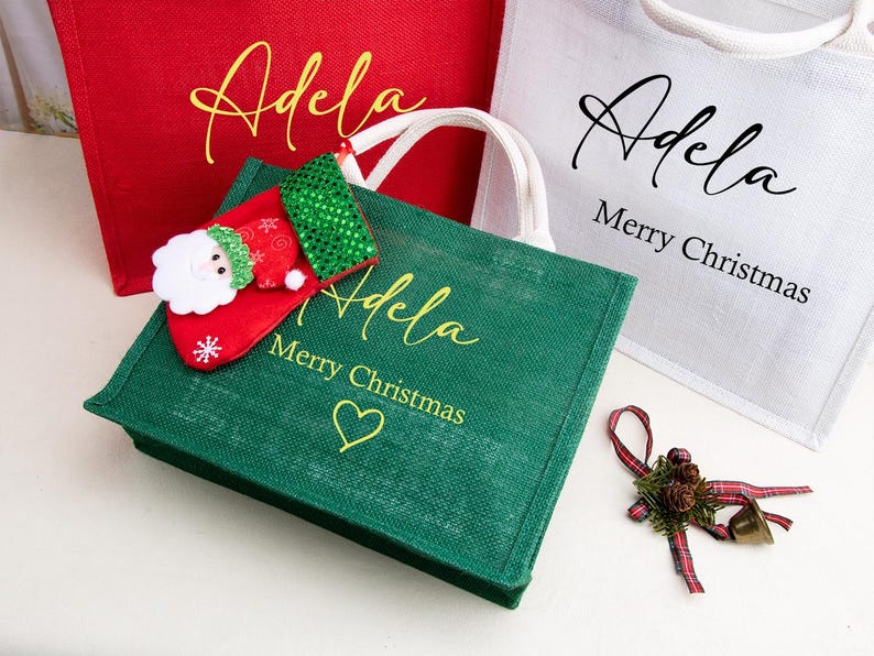 K&ouml;nnte beinhalten: Drei Jute-Tragetaschen in Rot, Gr&uuml;n und Wei&szlig; mit wei&szlig;en Henkeln. Die Taschen tragen den Namen Adela in Schreibschrift, dazu der Text Merry Christmas und ein Herzsymbol auf der gr&uuml;nen Tasche. Ein kleiner Weihnachtsstiefel und eine Schleife liegen bei.