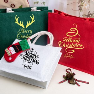 K&ouml;nnte beinhalten: Drei rechteckige Tragetaschen aus strukturiertem Stoff in Gr&uuml;n, Rot und Wei&szlig;. Die gr&uuml;ne Tasche zeigt ein gelbes Rentiergeweih und den Text Merry Christmas. Die rote Tasche hat ein gelbes, geschwungenes Baummotiv mit Merry Christmas und Adela. Die wei&szlig;e Tasche zeigt einen schwarzen Merry Christmas und Faith Schriftzug mit einem kleinen rot-gr&uuml;nen Schneemann-Strumpf-Anh&auml;nger am Griff.