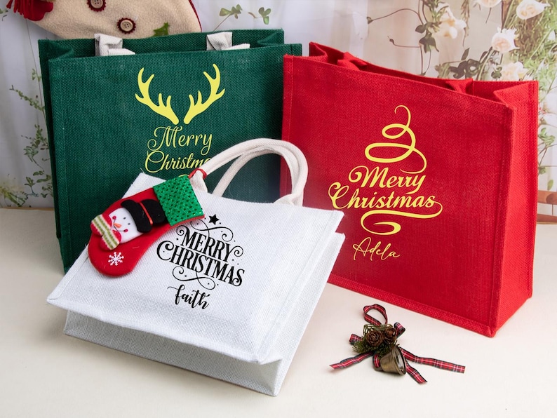 K&ouml;nnte beinhalten: Drei rechteckige Tragetaschen aus strukturiertem Stoff in Gr&uuml;n, Rot und Wei&szlig;. Die gr&uuml;ne Tasche zeigt ein gelbes Rentiergeweih und den Text Merry Christmas. Die rote Tasche hat ein gelbes, geschwungenes Baummotiv mit Merry Christmas und Adela. Die wei&szlig;e Tasche zeigt einen schwarzen Merry Christmas und Faith Schriftzug mit einem kleinen rot-gr&uuml;nen Schneemann-Strumpf-Anh&auml;nger am Griff.