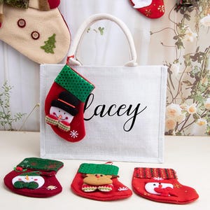 K&ouml;nnte beinhalten: Eine wei&szlig;e rechteckige Tragetasche mit dem Namen Lacey in schwarzer Schreibschrift. Ein kleiner roter Weihnachtsstrumpf mit gr&uuml;n gepunktetem Umschlag und Schneemann-Applikation h&auml;ngt am Griff. Drei weitere kleine Str&uuml;mpfe liegen davor.