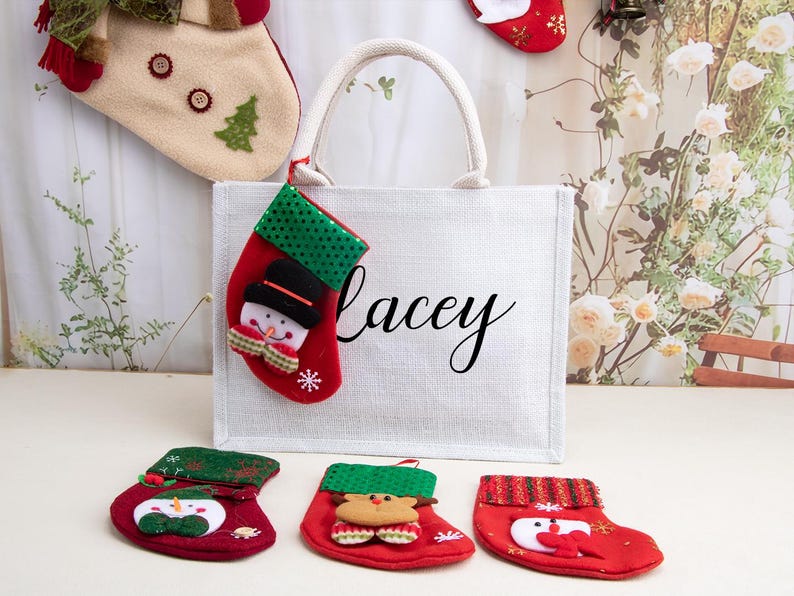 K&ouml;nnte beinhalten: Eine wei&szlig;e rechteckige Tragetasche mit dem Namen Lacey in schwarzer Schreibschrift. Ein kleiner roter Weihnachtsstrumpf mit gr&uuml;n gepunktetem Umschlag und Schneemann-Applikation h&auml;ngt am Griff. Drei weitere kleine Str&uuml;mpfe liegen davor.