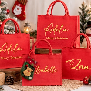 K&ouml;nnte beinhalten: Vier rote, rechteckige Tragetaschen mit jeweils zwei Henkeln. Drei Taschen zeigen goldene, kursive Namen und den Text Merry Christmas. Eine Tasche tr&auml;gt den goldenen Namen Isabelle und einen kleinen gr&uuml;nen Weihnachtsstrumpf mit Rentier.