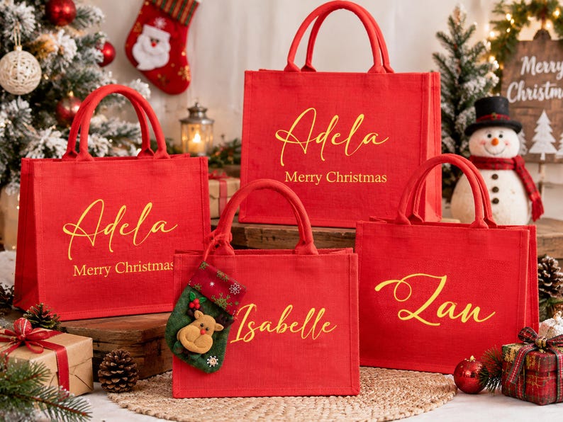 K&ouml;nnte beinhalten: Vier rote, rechteckige Tragetaschen mit jeweils zwei Henkeln. Drei Taschen zeigen goldene, kursive Namen und den Text Merry Christmas. Eine Tasche tr&auml;gt den goldenen Namen Isabelle und einen kleinen gr&uuml;nen Weihnachtsstrumpf mit Rentier.