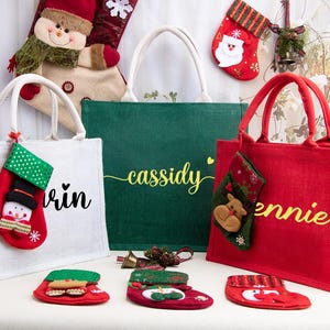 K&ouml;nnte beinhalten: Drei rechteckige Jute-Tragetaschen in Wei&szlig;, Gr&uuml;n und Rot mit wei&szlig;en Kordelgriffen und personalisierten Namen in Schreibschrift. Die Taschen sind mit kleinen h&auml;ngenden Weihnachtsstr&uuml;mpfen verziert, die Schneemann- und Rentier-Motive zeigen.