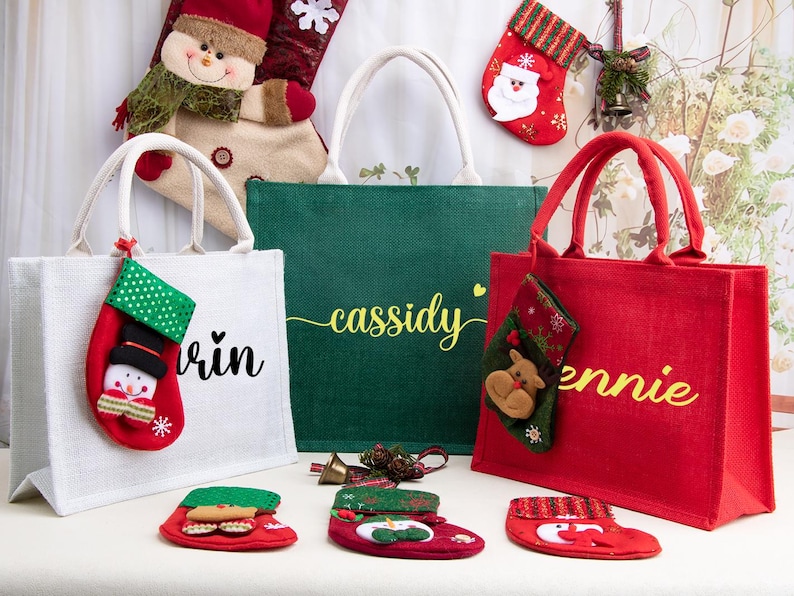 K&ouml;nnte beinhalten: Drei rechteckige Jute-Tragetaschen in Wei&szlig;, Gr&uuml;n und Rot mit wei&szlig;en Kordelgriffen und personalisierten Namen in Schreibschrift. Die Taschen sind mit kleinen h&auml;ngenden Weihnachtsstr&uuml;mpfen verziert, die Schneemann- und Rentier-Motive zeigen.