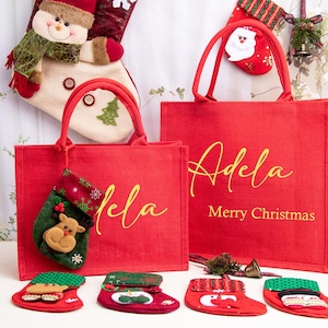 K&ouml;nnte beinhalten: Zwei rote, rechteckige Tragetaschen mit roten Henkeln, die jeweils den Namen Adela in goldener Schreibschrift tragen. Die gr&ouml;&szlig;ere Tasche enth&auml;lt den Text Merry Christmas in Gold. Mehrere kleine dekorative Weihnachtsstr&uuml;mpfe liegen um die Taschen.
