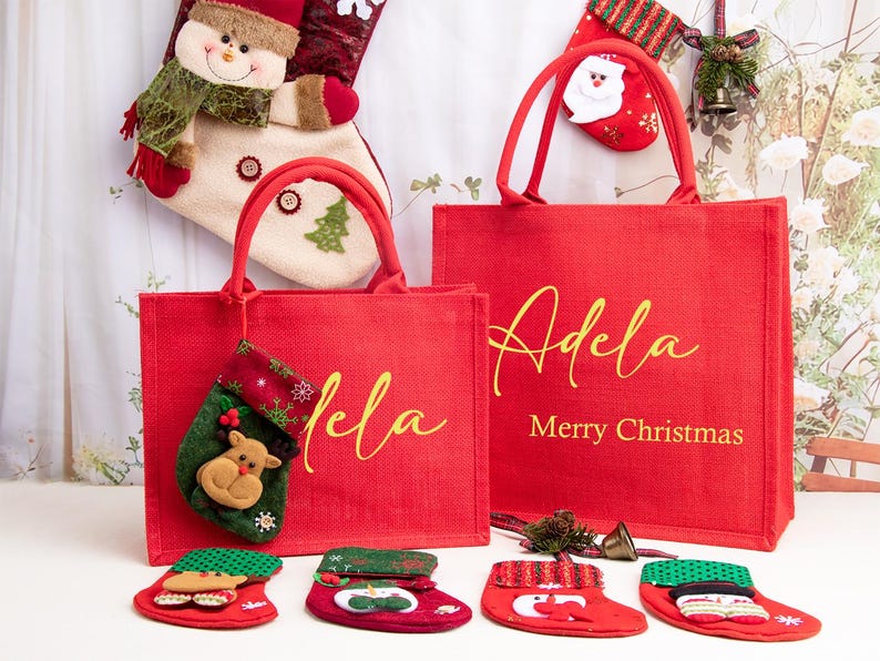 K&ouml;nnte beinhalten: Zwei rote, rechteckige Tragetaschen mit roten Henkeln, die jeweils den Namen Adela in goldener Schreibschrift tragen. Die gr&ouml;&szlig;ere Tasche enth&auml;lt den Text Merry Christmas in Gold. Mehrere kleine dekorative Weihnachtsstr&uuml;mpfe liegen um die Taschen.
