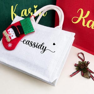 K&ouml;nnte beinhalten: Eine wei&szlig;e Jutetragetasche mit zwei cremefarbenen Henkeln, die den Namen "cassidy" in schwarzer Schreibschrift tr&auml;gt und mit einem kleinen, rot-gr&uuml;nen Filz-Weihnachtsstrumpf-Anh&auml;nger am Henkel versehen ist. Die Tasche liegt neben gr&uuml;nen und roten Taschen.