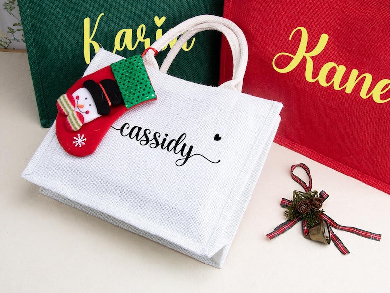 K&ouml;nnte beinhalten: Eine wei&szlig;e Jutetragetasche mit zwei cremefarbenen Henkeln, die den Namen "cassidy" in schwarzer Schreibschrift tr&auml;gt und mit einem kleinen, rot-gr&uuml;nen Filz-Weihnachtsstrumpf-Anh&auml;nger am Henkel versehen ist. Die Tasche liegt neben gr&uuml;nen und roten Taschen.