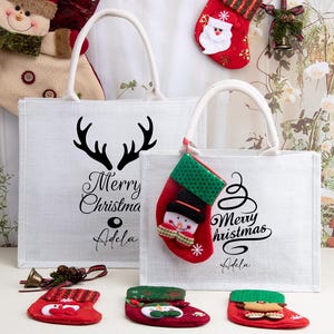 K&ouml;nnte beinhalten: Zwei wei&szlig;e, rechteckige Tragetaschen mit wei&szlig;en Henkeln, jeweils mit dem schwarzen Schriftzug Merry Christmas und dem Namen Adela bedruckt, wobei die gr&ouml;&szlig;ere Tasche ein schwarzes Rentiergeweih-Motiv und die kleinere einen roten Sockenanh&auml;nger hat.