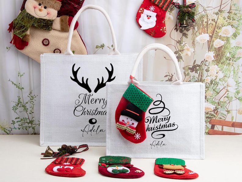 K&ouml;nnte beinhalten: Zwei wei&szlig;e, rechteckige Tragetaschen mit wei&szlig;en Henkeln, jeweils mit dem schwarzen Schriftzug Merry Christmas und dem Namen Adela bedruckt, wobei die gr&ouml;&szlig;ere Tasche ein schwarzes Rentiergeweih-Motiv und die kleinere einen roten Sockenanh&auml;nger hat.
