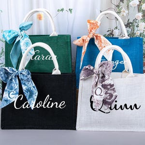 Puede incluir: Cuatro bolsas de mano personalizadas con acentos de tela y cinta de diferentes colores. Las bolsas están hechas de yute natural y tienen los nombres "Kara", "Caroline", "Quinn" y "Ange" impresos en ellas.