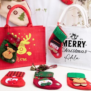 K&ouml;nnte beinhalten: Eine rote Jute-Tragetasche mit gelbem Rentier-Motiv und dem Namen Vera sowie eine wei&szlig;e Jute-Tasche mit dem Text Merry Christmas Adela. Beide Taschen haben kleine angeh&auml;ngte Str&uuml;mpfe, dazu liegen drei weitere Str&uuml;mpfe davor auf dem Untergrund.