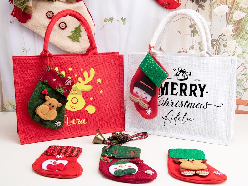 K&ouml;nnte beinhalten: Eine rote Jute-Tragetasche mit gelbem Rentier-Motiv und dem Namen Vera sowie eine wei&szlig;e Jute-Tasche mit dem Text Merry Christmas Adela. Beide Taschen haben kleine angeh&auml;ngte Str&uuml;mpfe, dazu liegen drei weitere Str&uuml;mpfe davor auf dem Untergrund.