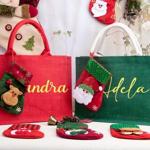 K&ouml;nnte beinhalten: Eine rote Jute-Tragetasche mit dem Namen Sandra und eine gr&uuml;ne Jute-Tragetasche mit dem Namen Adela. Jede Tasche hat einen kleinen h&auml;ngenden Weihnachtsstrumpf. Die rote Tasche hat einen Rentier-Strumpf und die gr&uuml;ne einen Weihnachtsmann-Strumpf.