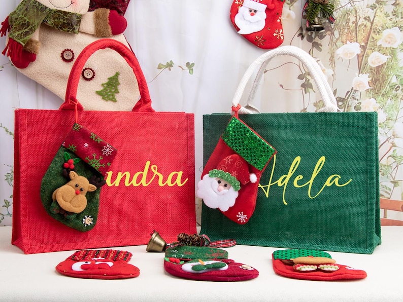 K&ouml;nnte beinhalten: Eine rote Jute-Tragetasche mit dem Namen Sandra und eine gr&uuml;ne Jute-Tragetasche mit dem Namen Adela. Jede Tasche hat einen kleinen h&auml;ngenden Weihnachtsstrumpf. Die rote Tasche hat einen Rentier-Strumpf und die gr&uuml;ne einen Weihnachtsmann-Strumpf.