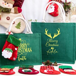 K&ouml;nnte beinhalten: Zwei gr&uuml;ne Jute-Tragetaschen mit wei&szlig;en Henkeln, jeweils mit gelbem Schriftzug Merry Christmas und personalisierten Namen. Die Taschen sind mit kleinen roten Stoffstr&uuml;mpfen verziert, darunter einer mit einem Weihnachtsmann-Gesicht.