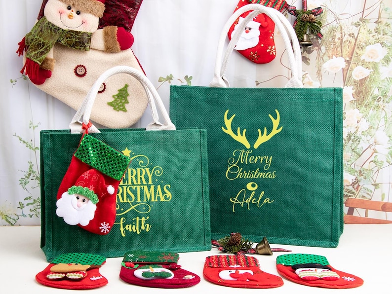 K&ouml;nnte beinhalten: Zwei gr&uuml;ne Jute-Tragetaschen mit wei&szlig;en Henkeln, jeweils mit gelbem Schriftzug Merry Christmas und personalisierten Namen. Die Taschen sind mit kleinen roten Stoffstr&uuml;mpfen verziert, darunter einer mit einem Weihnachtsmann-Gesicht.
