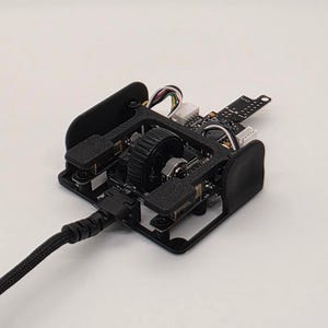 Può includere: Un assemblaggio interno di mouse per computer nero con rotellina di scorrimento centrale, circuito stampato a vista e cavo intrecciato nero. Il dispositivo include un telaio in plastica nera, due pulsanti rettangolari e un connettore bianco.