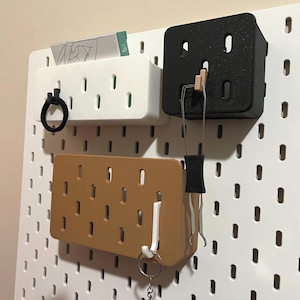 May include: A white pegboard with three attached storage containers in white, black, and brown. The containers have various items hanging from them, including a black hair tie, a wooden clothespin, and a key ring.