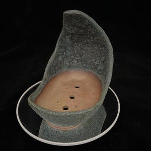 May include: A handcrafted bonsai pot with a textured, grey-green exterior and a light brown interior. The pot has a distinctive, organic shape and three drainage holes. It sits on a clear, round saucer.