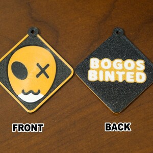 May include: A pair of diamond-shaped keychains. One side features a yellow alien face with a black eye and an X for a mouth. The other side has the text "BOGOS BINTED" in yellow. Both are outlined in black.