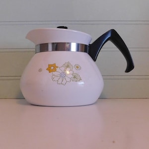 May include: White vintage coffee pot with a black handle and lid. The pot has a silver band around the middle and is decorated with yellow and green floral designs. The coffee pot is sitting on a white surface.