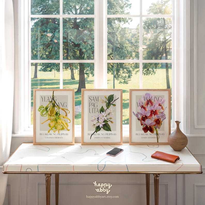 Może przedstawiać: Trzy oprawione botaniczne wydruki artystyczne z ilustracjami kwiat&oacute;w ylang-ylang, sampaguita i waling-waling. Wydruki są eksponowane na białym stole z dekoracyjnym wzorem, przed oknem. Na stole znajdują się r&oacute;wnież mały brązowy wazon, telefon i portfel.