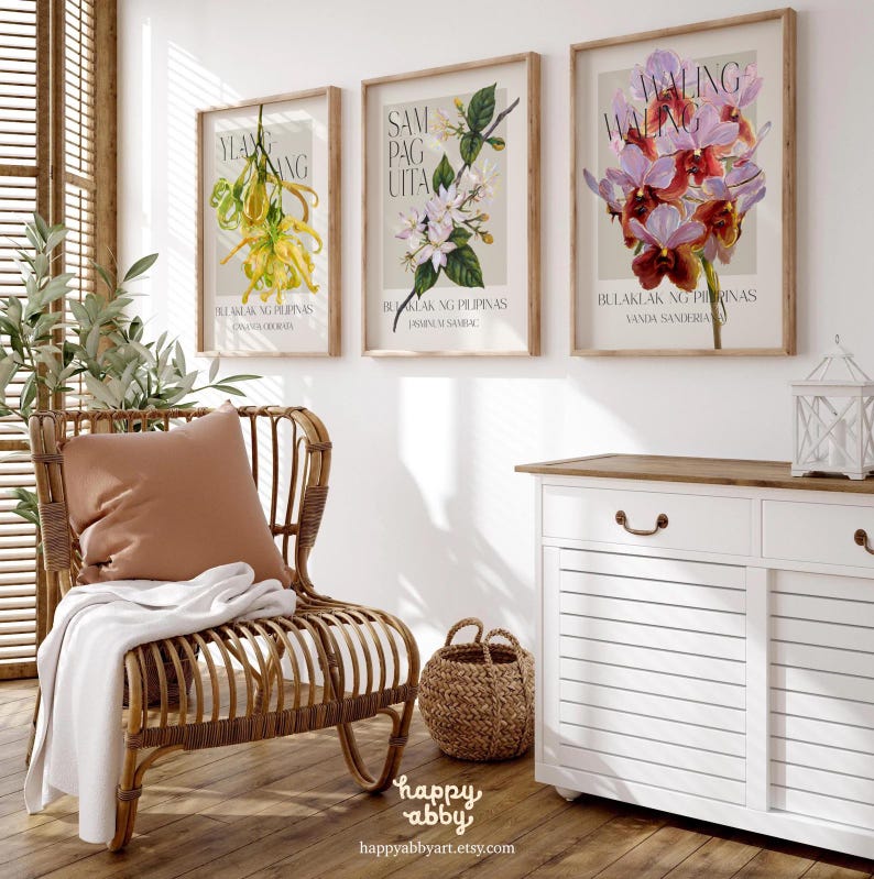 Może przedstawiać: Trzy oprawione grafiki botaniczne z ilustracjami kwiatowymi i tekstem w języku obcym. Grafiki są w drewnianych ramach i wiszą na białej ścianie. Na pierwszym planie znajduje się wiklinowy fotel z poduszką i kocem, pleciony kosz i biała szafka.
