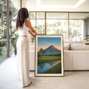May include: A framed landscape art print featuring a volcano at sunset, with a river and palm trees in the foreground. The artwork has a black frame and is displayed indoors. The print has the text "happy abby" and "happyabbyart.etsy.com".