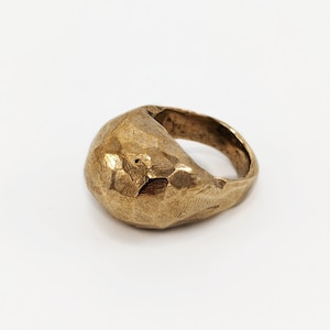 May include: A gold-colored ring with a textured, hammered surface. The ring has a rounded, irregular shape and a thick band. The ring is set against a white background.
