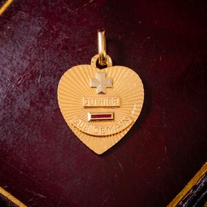May include: A gold heart-shaped pendant featuring a sunburst texture, a small cross, and the text QU'HIER. Below the text is a horizontal bar with three inset red rubies, followed by the text QUE DEMAIN, all centered on the pendant surface.