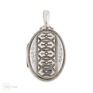 Op de afbeelding: Ovale zilveren locket met ingewikkelde gegraveerde patronen. De locket heeft een centraal geometrisch ontwerp en delicate bloemaccenten langs de randen. Bovenop zit een kleine lus voor een ketting. Het is een vintage stuk.