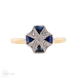 May include: A gold ring featuring a cross-shaped centrepiece set with four triangular blue sapphires and small clear stones. The central setting is crafted from a silver-toned metal with intricate detailing, contrasting with the smooth gold band.