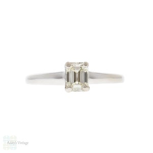 May include: A simple solitaire engagement ring with a square emerald cut diamond set in a thin white gold band.