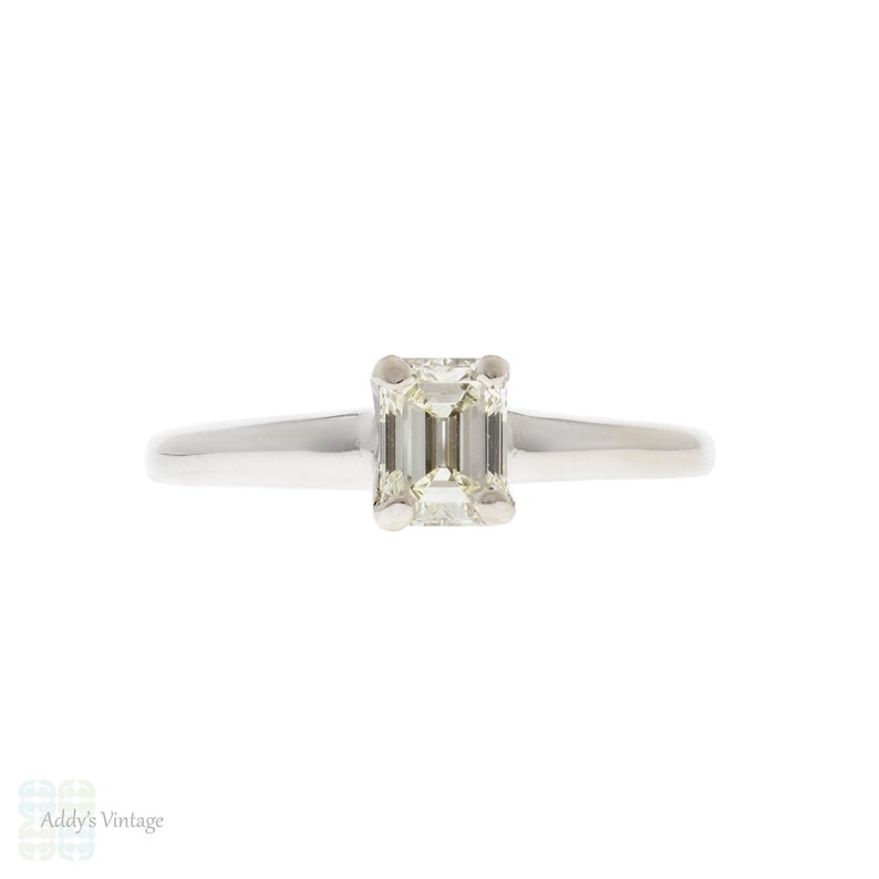 May include: A simple solitaire engagement ring with a square emerald cut diamond set in a thin white gold band.