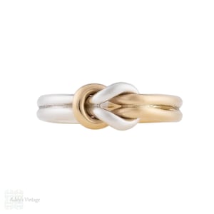 Puede incluir: Un anillo de dos tonos con un diseño de nudo. El anillo tiene un centro de nudo de color dorado y bandas de color plateado. El anillo es una toma de primer plano sobre un fondo blanco.
