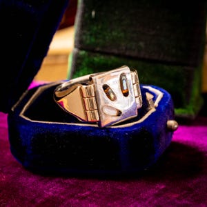 May include: A gold ring featuring a rectangular face with three inlaid iridescent shell accents. The ring has a polished metal band and a hinged-style design on the sides of the setting, showcasing a smooth, reflective surface with natural shell details.