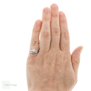 May include: A close-up of a hand wearing a silver engagement ring with a square-shaped center stone and a matching wedding band with smaller stones.
