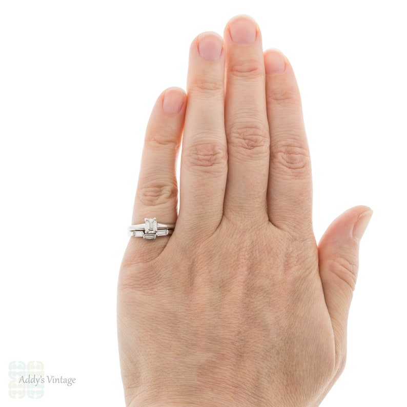 May include: A close-up of a hand wearing a silver engagement ring with a square-shaped center stone and a matching wedding band with smaller stones.