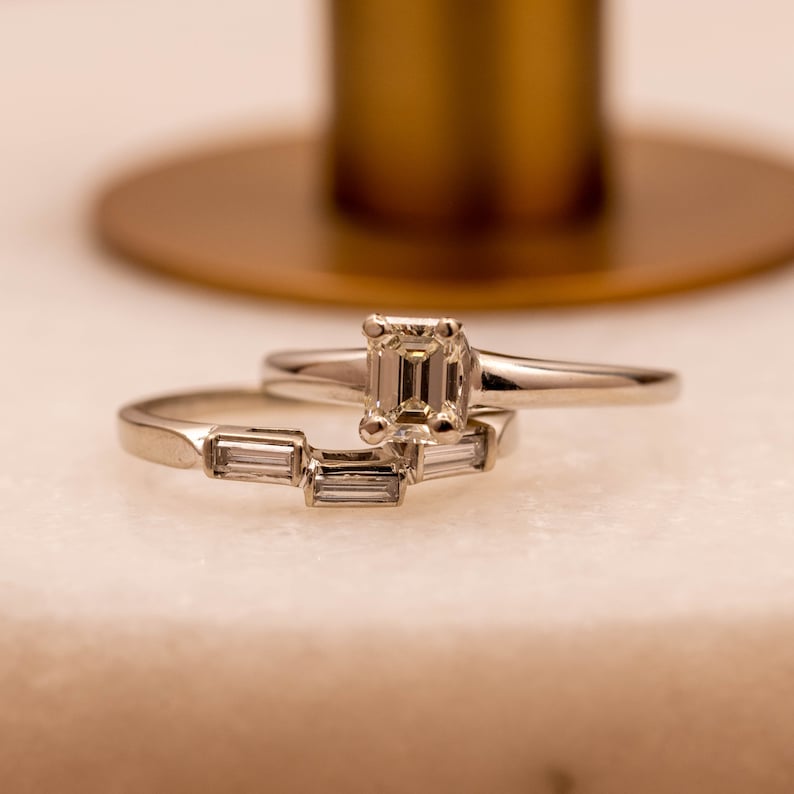 May include: Two silver rings with diamonds. The top ring features a larger, emerald-cut diamond. The bottom ring has three smaller, baguette-cut diamonds.
