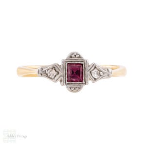May include: Vintage gold ring with a square, deep red gemstone at the center, flanked by two smaller diamonds. The ring has a detailed, ornate design with a gold band and silver-colored accents around the gemstones.