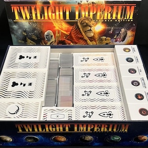 Puede incluir: Caja de juego de mesa abierta titulada "Twilight Imperium Fourth Edition". Contiene piezas de juego blancas, cartas y fichas de facción. La ilustración de la caja presenta una figura de león y escenas espaciales.