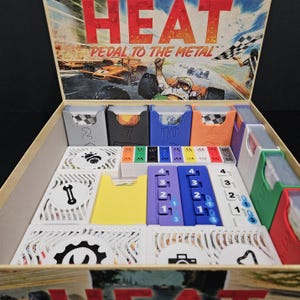 Może przedstawiać: Otwarty box gry planszowej zatytułowany "HEAT Pedal to the Metal". Pudełko zawiera różne elementy gry, w tym numerowane uchwyty na karty w kolorze szarym, czarnym, niebieskim i pomarańczowym. Są też karty i kolorowe pionki.