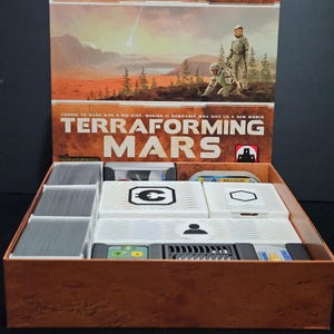 May include: Open board game box titled "Terraforming Mars." The box features a Martian landscape illustration with two figures in space suits. Inside, white game components are visible, including labeled boxes and card holders. The box is brown with the game title prominently displayed.