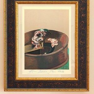 May include: Framed print of a Francis Bacon painting. The artwork depicts a figure in a circular structure with a dark brown frame and gold accents. The print has a beige background and a signature at the bottom.