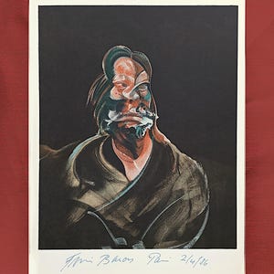 May include: A framed print of a portrait with a dark backdrop. The artwork shows a distorted face in shades of orange, green, and white. The print is signed and dated "2/4/86" in blue ink. The frame is white, and the background is a deep red.