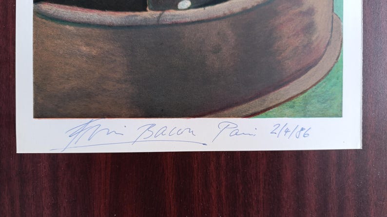 May include: A framed artwork featuring a brown, bowl-shaped object with a white rim, set against a green and brown background. The artist's signature and the date 2/4/86 are visible in blue ink.