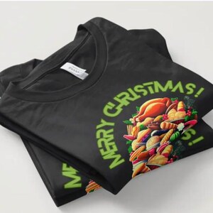 May include: Black t-shirt with a round neck, neatly folded. The shirt displays a green "MERRY CHRISTMAS!" design and a colourful illustration of a festive meal, including a roasted turkey and various side dishes.