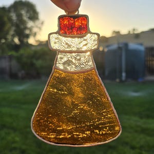 May include: Stained glass pendant in a bottle shape, featuring a copper frame. The top is red, the middle is clear with a floral design, and the bottom is amber. The pendant is held up against a green lawn and a sunset.